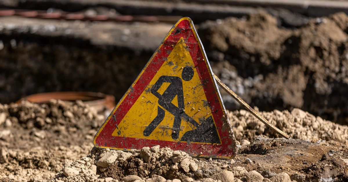 A weathered roadworks sign shows a person digging with a shovel. The sign is partially buried in a pile of dirt.