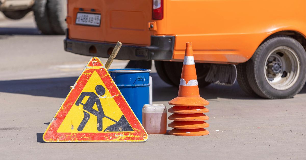 A worn roadwork sign and a stack of orange cones sit beside a bright orange utility van on pavement.